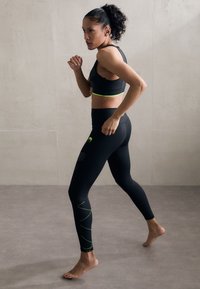 Črn športni set z športnim nedrčkom z racerback rezom, ki ima zeleno zaplato, in visokim pasom leggings z teksturiranimi vzorci in neon zelenimi črtami.