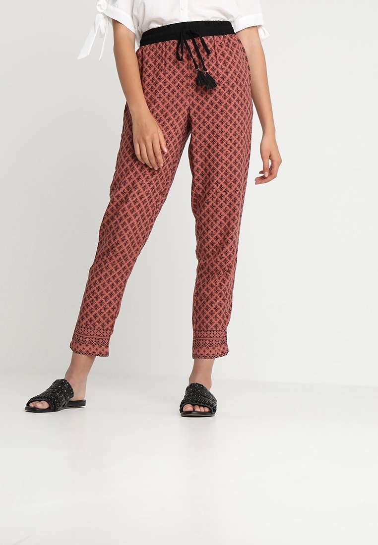 Pantalons à motifs rouges avec une taille élastique noire et un cordon de serrage, présentant des designs complexes et des jambes fuselées. Sandales noires à enfiler.