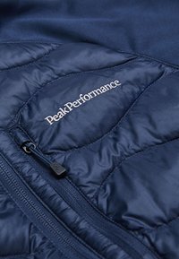 Marineblaue gesteppte Jacke aus weichem Material, mit einem durchgehenden Reißverschluss und einem weißen gestickten Logo, sowie einer glatten Textur und einem eleganten Design.