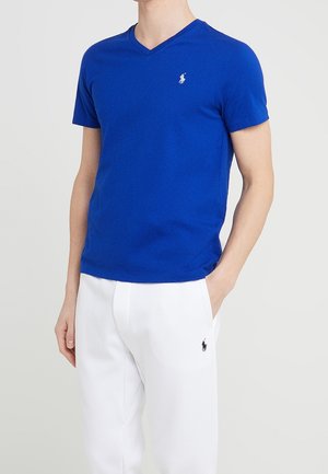 Hombre con camiseta azul de cuello en V y pantalones blancos con logotipos en el pecho y el muslo, de pie con una mano en el bolsillo.