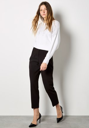 Kobieta ubrana w biały, długi, perforowany sweter, czarne, krótkie spodnie z wysokim stanem oraz czarne, szpiczaste szpilki, stojąca przy białej ścianie.