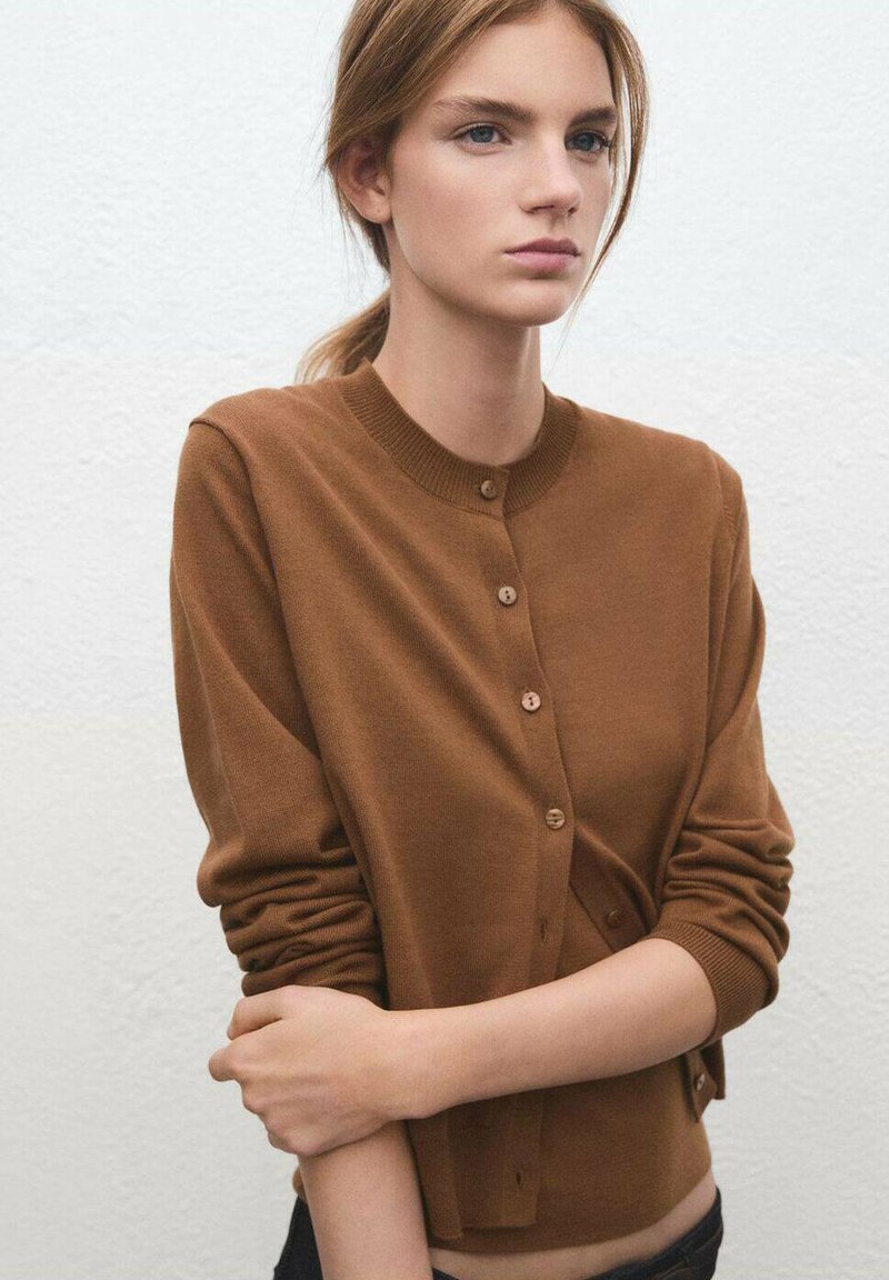 Cárdigan de punto marrón con escote redondo, botones de madera y mangas largas, exhibido sobre un fondo texturizado en tonos neutros.