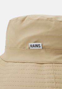 Cappello a secchiello beige con una superficie liscia e impermeabile, caratterizzato da cuciture orizzontali e un'etichetta bianca con il nome del marchio "RAINS."