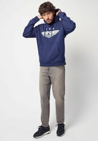 Felpa navy con tasca frontale e stampa grafica che presenta ali e testo; abbinata a jeans grigi chiari e scarpe sportive nere.