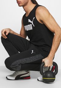 Camisola tipo tank top preta da Puma com logo branco; calças pretas com detalhes cinzentos; segurando um kettlebell preto; usando ténis atléticos cinzentos e vermelhos.