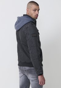 Svart vadderad jacka med en blå huva. Den har dragkedja och knapplås, ribbade ärmslut och ett emblem på vänster ärm. Grå jeans kompletterar looken.