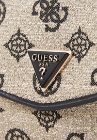 Béžová taška z texturované látky s černými květinovými vzory, ozdobená trojúhelníkovým černo-zlatým logem "GUESS USA". Měkký, strukturovaný tvar s koženými detaily.