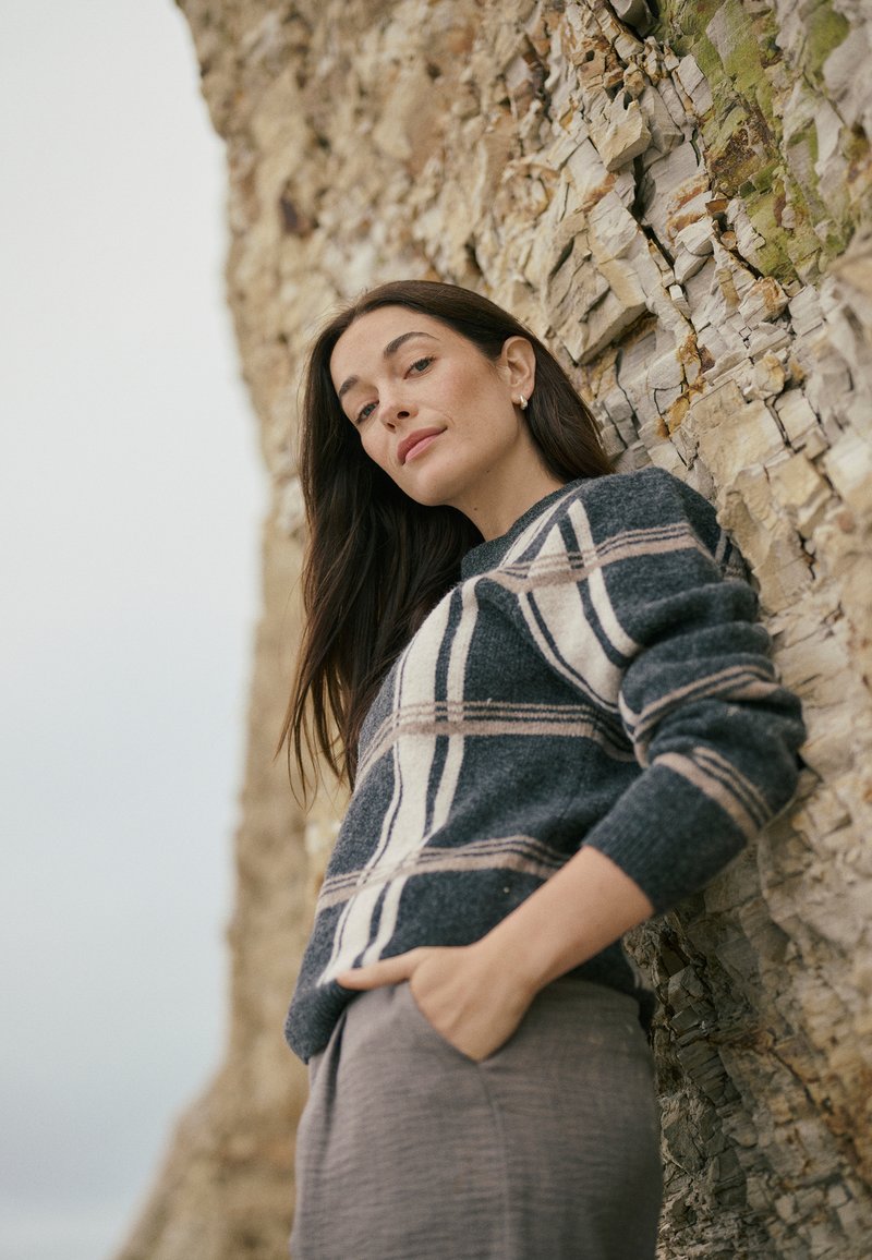 Grauer Wollpullover mit horizontalen blauen und cremefarbenen Streifen, mit langen Ärmeln und lockerer Passform, kombiniert mit einem grauen Rock aus strukturiertem Stoff.