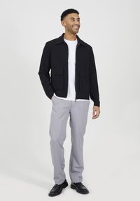 Giacca nera con zip e due tasche frontali, indossata sopra una maglietta bianca, abbinata a pantaloni grigio chiaro e scarpe nere, in piedi contro uno sfondo semplice.