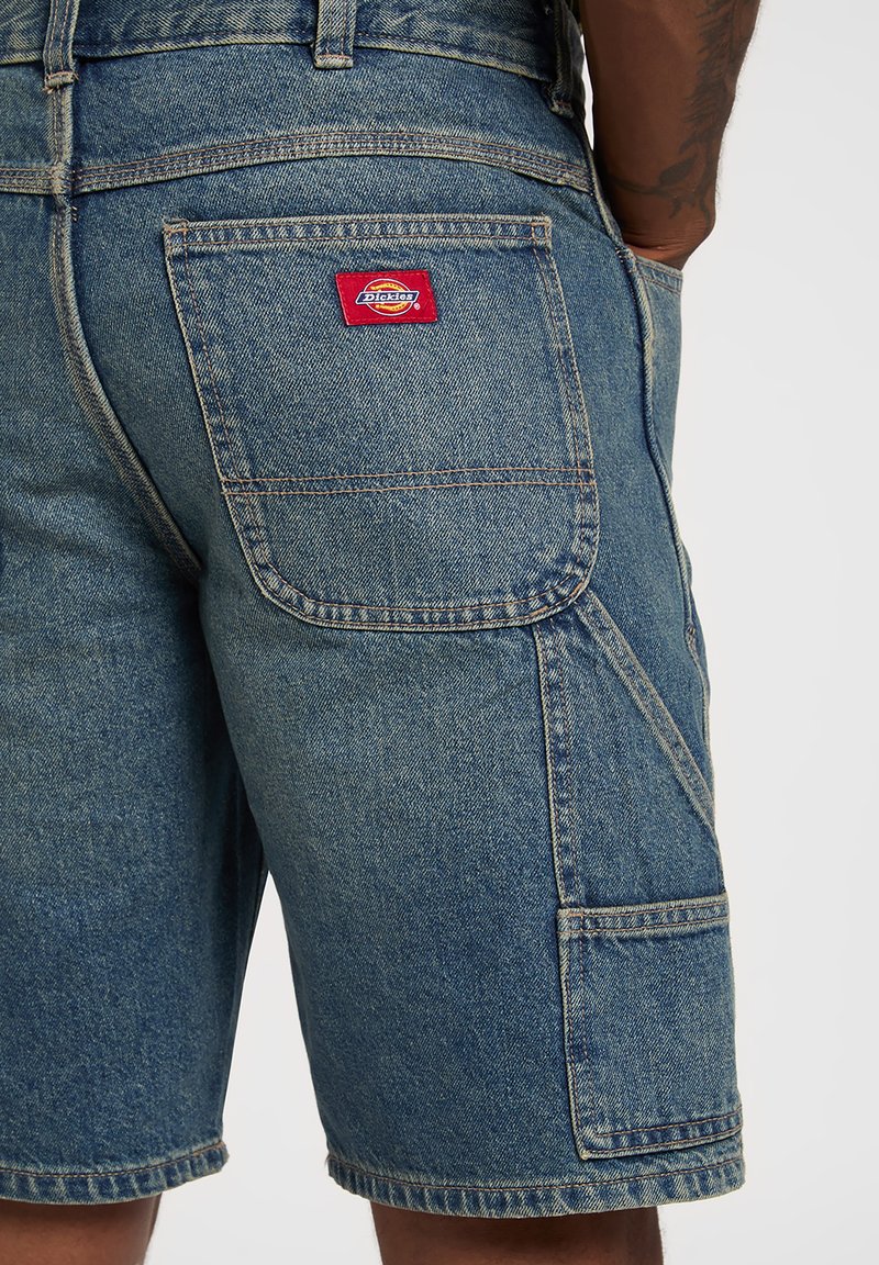 Pantaloncini di denim con una tasca posteriore decorata da una toppa con il logo rosso Dickies su una persona con un tatuaggio visibile sul braccio e la mano in tasca.