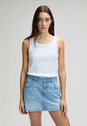 Giovane donna con capelli lunghi e scuri che indossa una canottiera bianca e una minigonna di jeans azzurro chiaro contro uno sfondo chiaro e uniforme.