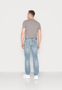T-shirt cinza de mangas curtas, jeans azuis desgastados, e ténis brancos. Os jeans têm um emblema de couro na parte de trás da cintura.