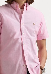 Ljusrosa kortärmad skjorta med knappar, tillverkad av tyg med en slät yta. Har en liten broderad logotyp på bröstet och vita knappar.