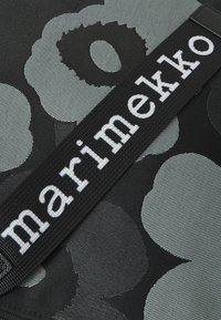 Black fabric with large gray floral patterns and a black strap displaying the word "marimekko" in white lettering. Textured surface.