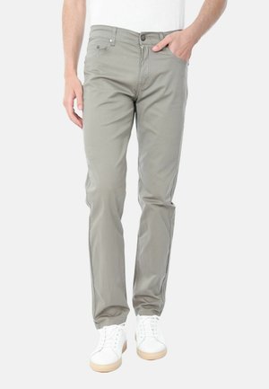 Uomo con pantaloni grigi chiaro a taglio slim, sneakers bianche e camicia bianca, con una mano in tasca, su sfondo semplice.