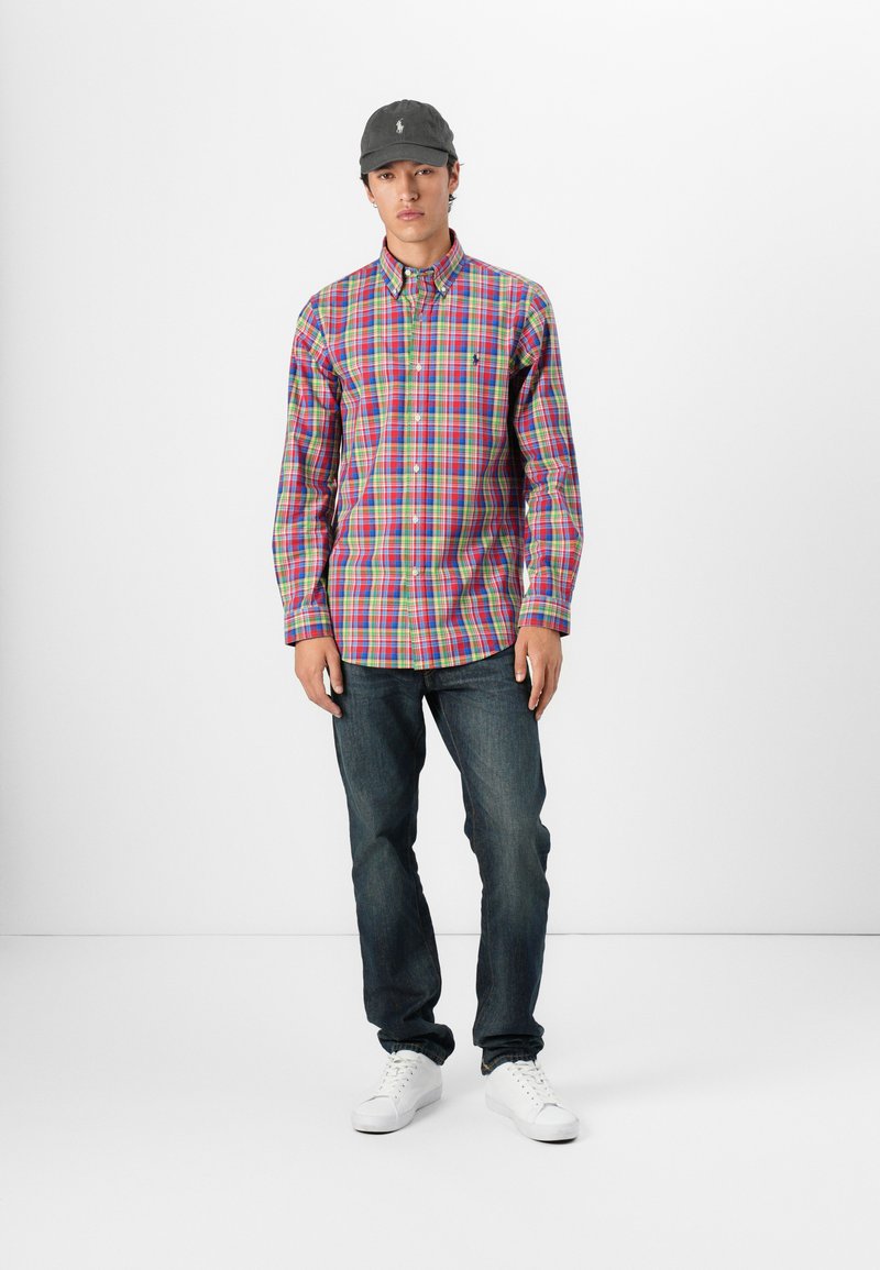 Męska koszula w kratę z guzikami w żywych kolorach, połączona z niebieskimi dżinsami i białymi sneakersami. Czapka jest szara z logo.