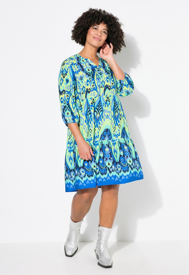 Vestido colorido a la altura de la rodilla con un estampado azul y verde, escote en V, mangas tres cuartos y una falda en capas. Combinado con botas de tobillo plateadas.