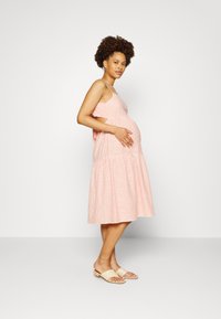 Vestido de maternidade listrado em rosa claro com saia em camadas, apresentando alças finas e laterais recortadas. Combinado com sandálias de slide em bege.