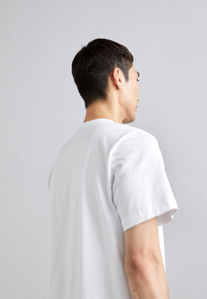 White cotton t-shirt with short sleeves and a crew neckline, displayed on a person from behind against a light gray background.