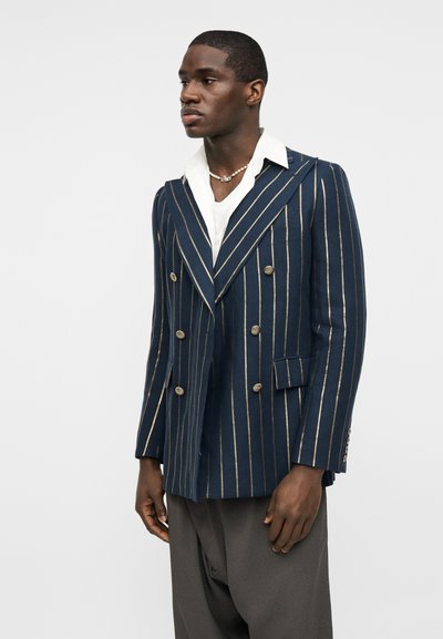 Άνδρας με μπλε σκούρο διπλό σακάκι με χρυσές κάθετες ρίγες, λευκό πουκάμισο, κολιέ με πέρλες και σκουρόχρωμο γκρι παντελόνι, στέκεται μπροστά σε ανοιχτό φόντο.