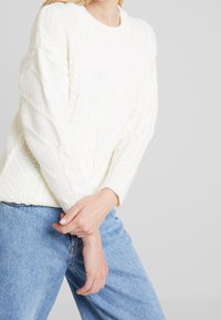 Femme portant un pull en laine torsadée blanc et un jean bleu clair, les mains jointes devant un fond gris uni.
