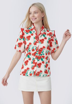 Jonge vrouw draagt een witte blouse met rode bloemenprint en ruches, gecombineerd met een witte minirok, glimlachend en poserend tegen een effen achtergrond.