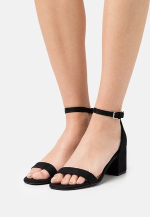 Černé semišové sandály na podpatku se širokými pásky, přezkou na kotníku a čtvercovým blokovým podpatkem. Otevřená špička ukazuje na nehty. Hladký povrch.
