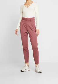 Pitkähiha cream-värinen paita, jossa on ristikkäinen kaula-aukko, yhdistetty korkeavyötäröisiin, räätälöityihin vaaleanpunaisiin housuihin ja vaaleisiin lenkkikenkiin.