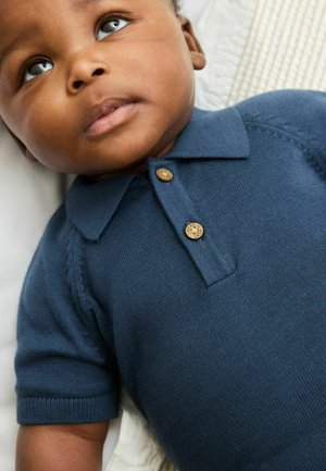 Sötét bőrű, kékes-szürke szemű baba fekszik, tengerészkék rövid ujjú kötött galléros pólót visel, fából készült gombokkal.