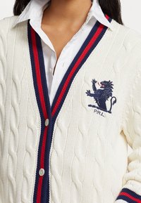 Kabelstickad cardigan i krämfärg med ribbade accentfärger i marinblått och rött. Har en broderad lion crest och knappstängning. Mjuk textur.