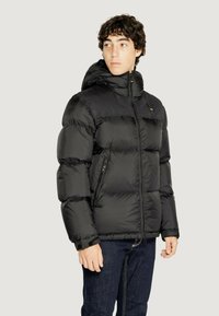 Giacca a vento nera con cappuccio, dotata di chiusura con zip, polsini a coste e numerose tasche, esposta su uno sfondo neutro.