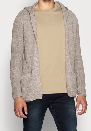 Mężczyzna w beżowej dzianinowej otwartej bluzie z kapturem, jasnobrązowym t-shircie i ciemnoniebieskich dżinsach, stojący na jasnym tle, ręce swobodnie wzdłuż ciała.