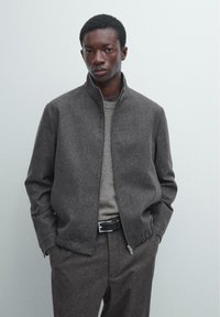 Graue Reißverschlussjacke mit Stehkragen aus strukturiertem Stoff. Kombiniert mit einem hellgrauen Pullover und maßgeschneiderten dunkelgrauen Hosen.