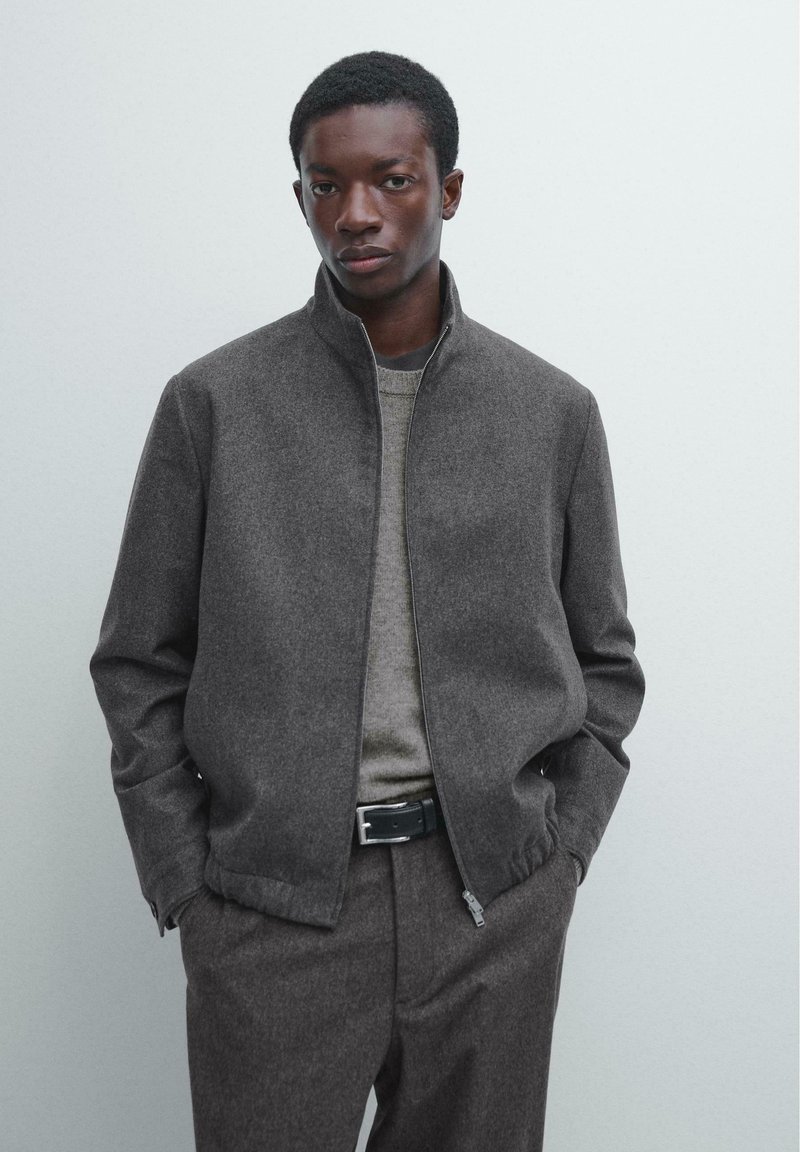 Graue Reißverschlussjacke mit Stehkragen aus strukturiertem Stoff. Kombiniert mit einem hellgrauen Pullover und maßgeschneiderten dunkelgrauen Hosen.