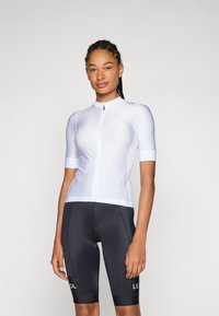 Weißes, geripptes Radtrikot mit kurzen Ärmeln und einem Reißverschluss auf der Vorderseite, kombiniert mit schwarzen Fahrradhosen mit weißer Markenkennzeichnung. Glatte Textur.