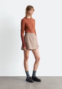Rostfarbenes Langarmoberteil, beiger Rock, graue Socken, glänzende schwarze Slipper. Minimalistisches Design, figurbetonte Silhouette, strukturierter Stoff, subtile Steppdetails.