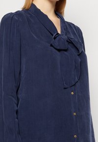 Granatowa bluzka z wiązaniem przy szyi, długimi rękawami, teksturowanym materiałem oraz złotymi guzikami na przodzie. Gładki, lekki materiał.