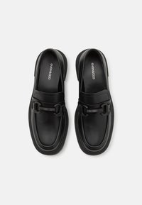 Schwarze Leder-Loafer mit glatter Textur, runder Form und dekorativem Kettenakzent über dem Spann. Minimalistisches Design mit leichtem Plateau.