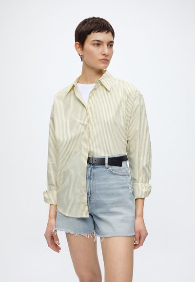 Femme aux cheveux courts portant une chemise jaune ample à rayures sur un top blanc, rentrée dans un short en denim taille haute avec une ceinture noire.