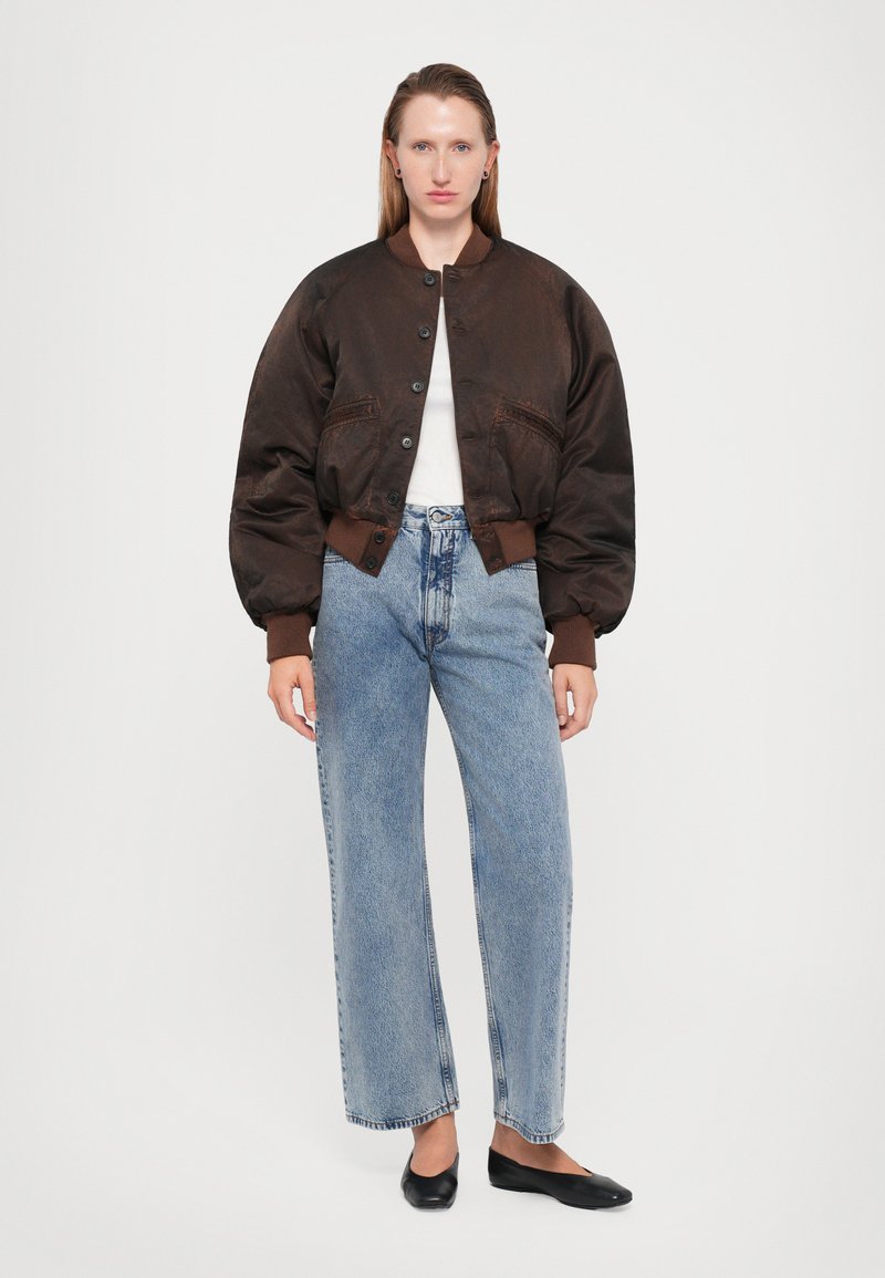 Veste aviateur marron avec fermeture éclair à l'avant, poignets côtelés, coupe oversize, associée à un jean taille haute bleu et des ballerines noires. Haut blanc visible.