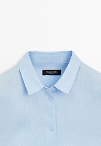 Lichtblauw overhemd met knopen en puntige kraag, gemaakt van linnen stof, met zichtbare halslijn en drie knopen.