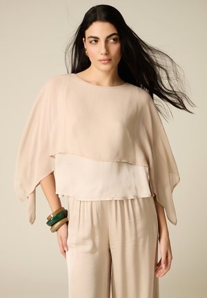 Donna con lunghi capelli neri che indossa un top beige ampio a strati e pantaloni abbinati, accessoriata con braccialetti verdi e marroni.