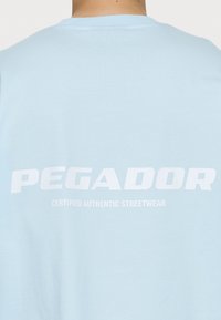 Svetlo modra bombažna majica s krojnim ovratnikom, ki ima na hrbtni strani velike belega logotipa "PEGADOR" in besedilo "CERTIFIED AUTHENTIC STREETWEAR".