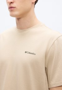 Μπεζ κοντομάνικη μπλούζα από μαλακό βαμβάκι, με μικρό πράσινο λογότυπο "Columbia" στην αριστερή πλευρά του στήθους. Ομαλή υφή και κλασική εφαρμογή.