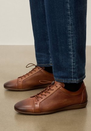 Hnedé kožené šnurovacie topánky s perforovanými stranami, nosené s tmavomodrými džínami na béžovej podlahe.