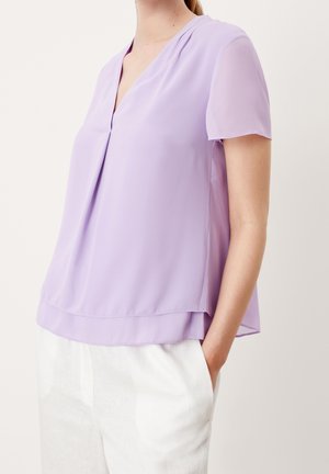 Femme portant un chemisier lavande ample à manches courtes avec un col en V et un pantalon blanc, debout avec une main dans la poche.