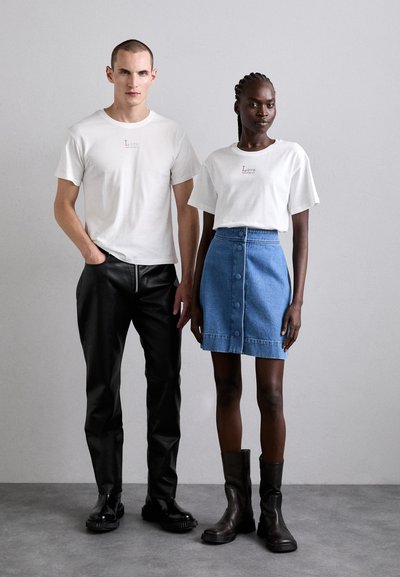 Două modele stau pe un fundal neutru. Modelul masculin poartă un tricou alb și pantaloni din piele neagră. Modelul feminin poartă un tricou alb și o fustă din denim albastru cu nasturi. Amândouă poartă încălțăminte neagră.