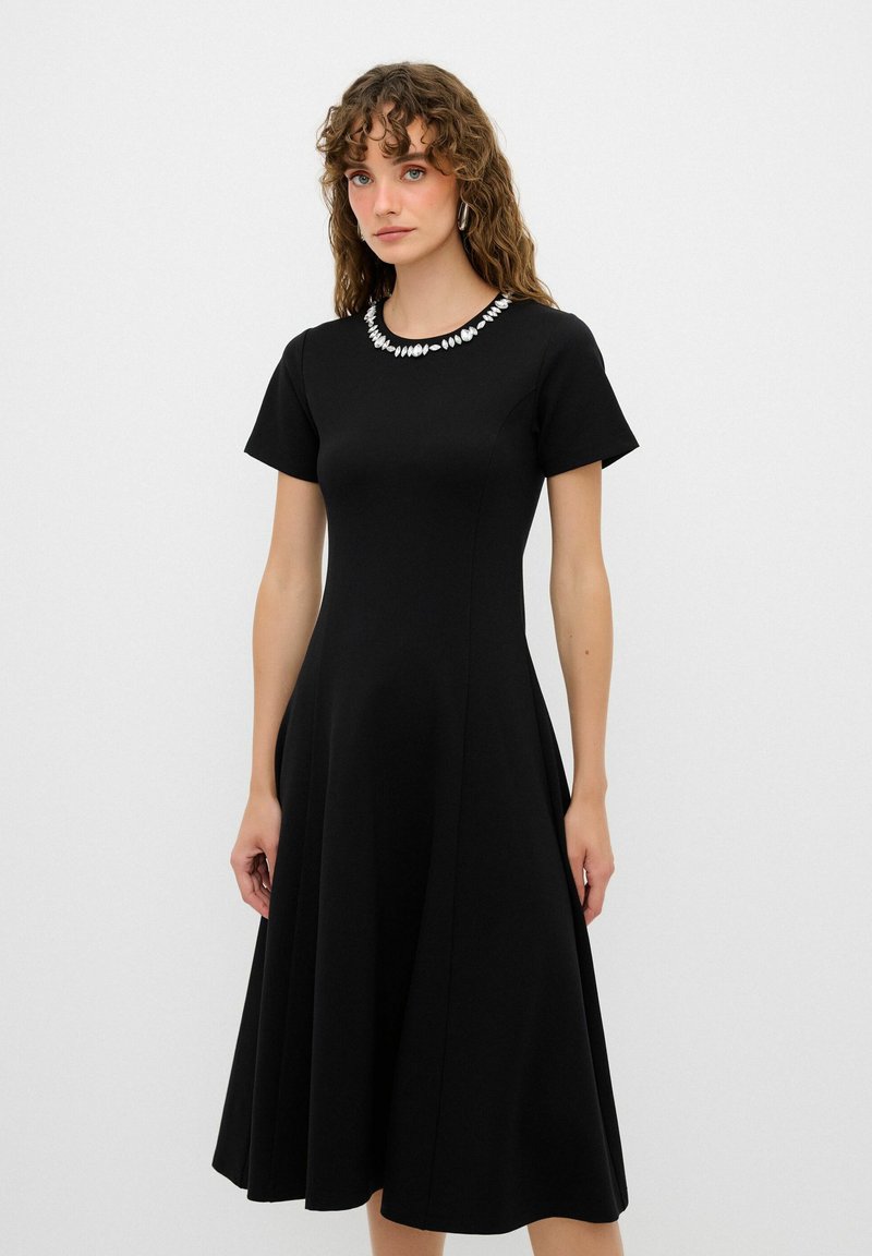 Schwarzes Kurzarmkleid mit weitem Rock, verziert mit strukturierten weißen Perlen um den Ausschnitt. Glatter Stofffinish.