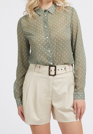Femme portant un chemisier transparent vert à pois avec des boutons, rentré dans un short beige taille haute avec une ceinture assortie et une boucle carrée.