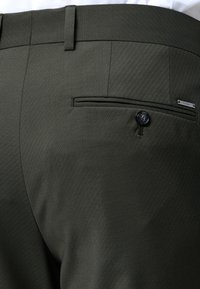 Pantalon vert foncé sur mesure avec des rayures diagonales discrètes, doté d'une poche arrière boutonnée et d'une petite étiquette métallique de la marque.